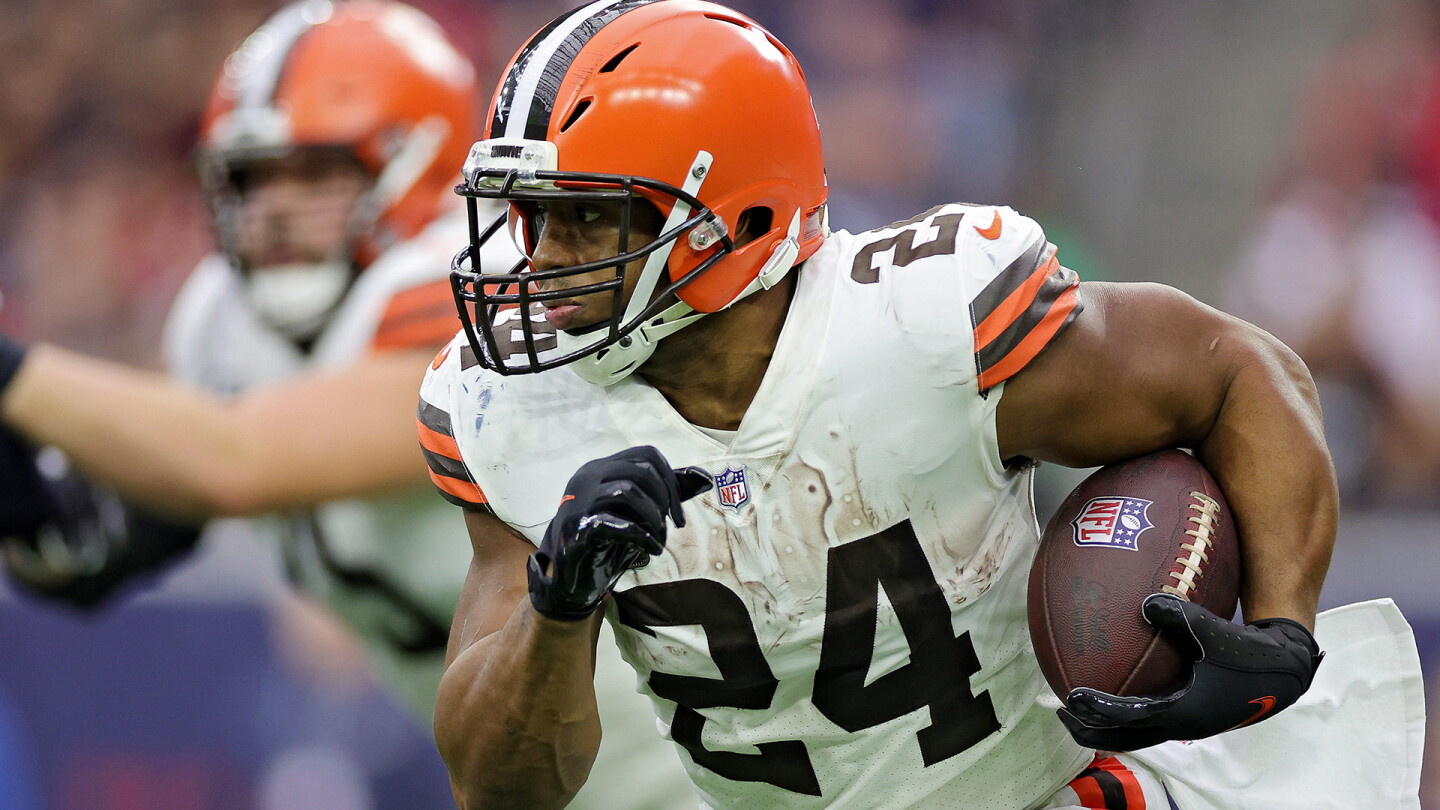 Chubb in a press conference, microphones in front, serious expression, Browns backdrop, reporters taking notes.